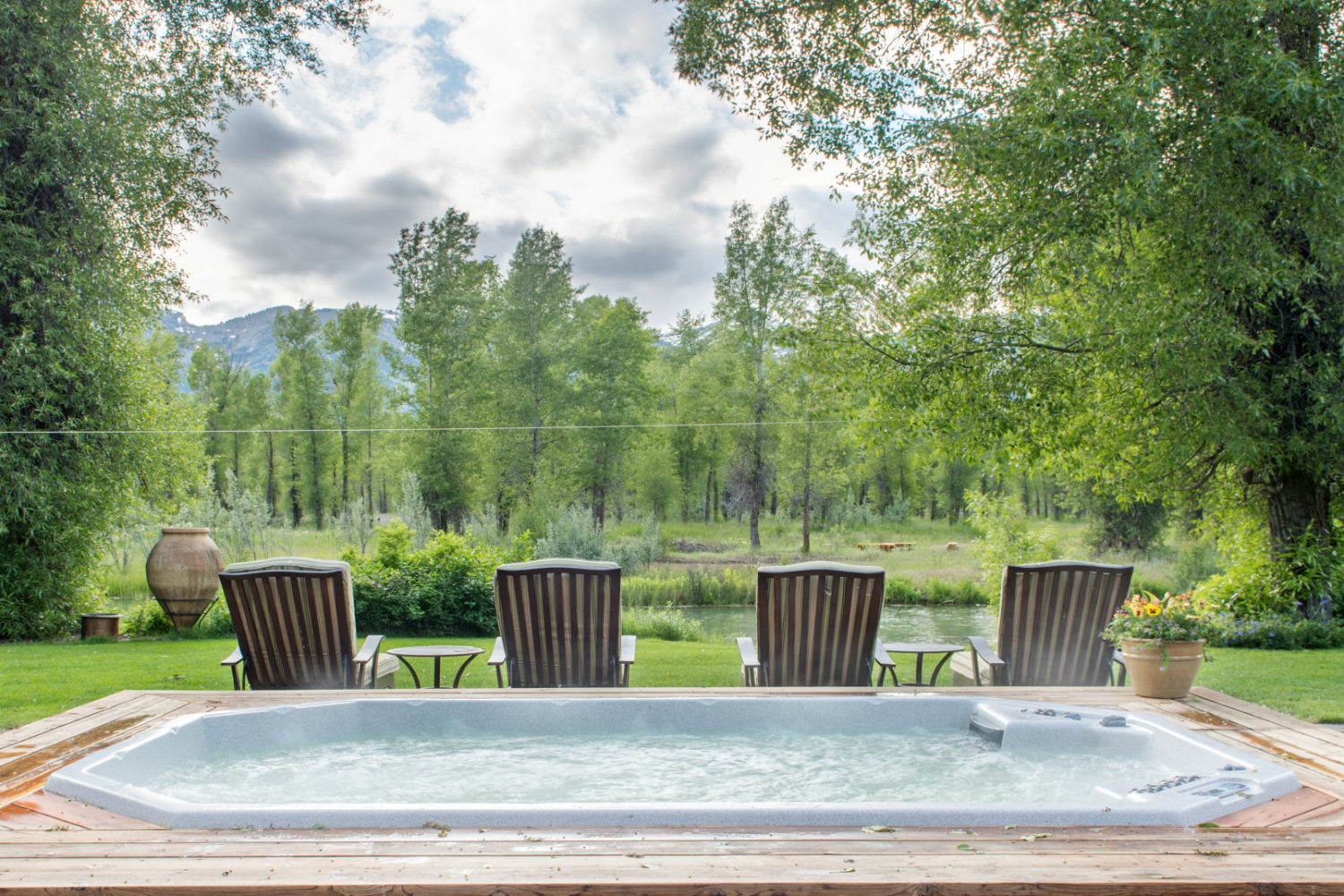 A large hot tub and 4 lawn chairs facing a creek, trees, and mountains