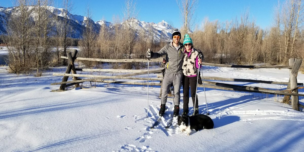 A couple pose in front of the mountains while cross country skiing with their dog