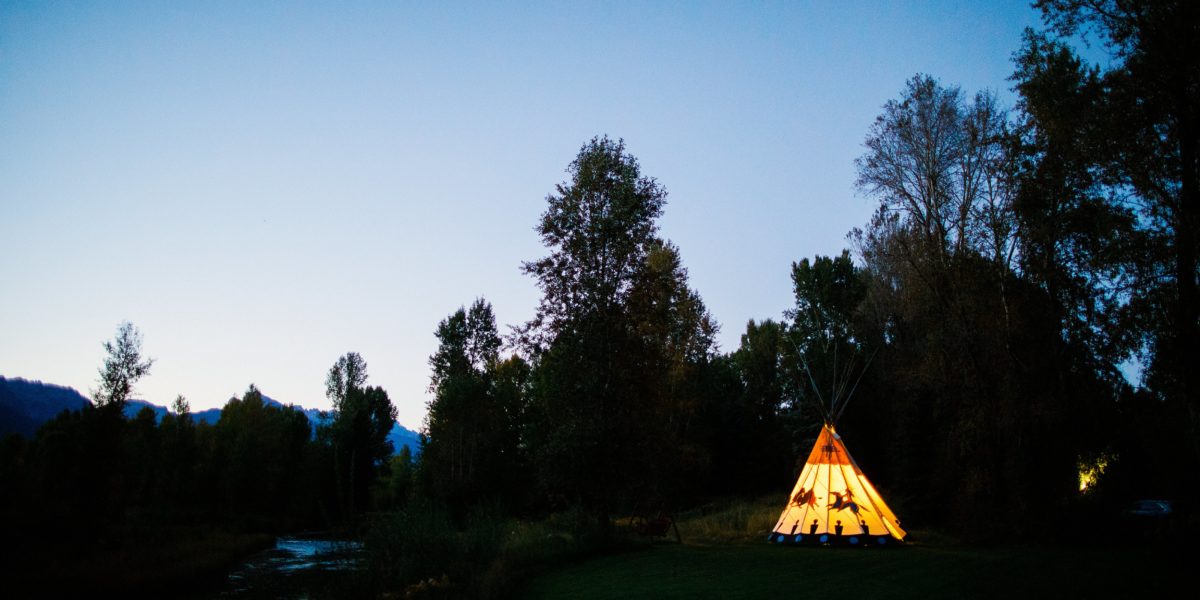A dusk image of mountains and trees with a glowing teepee