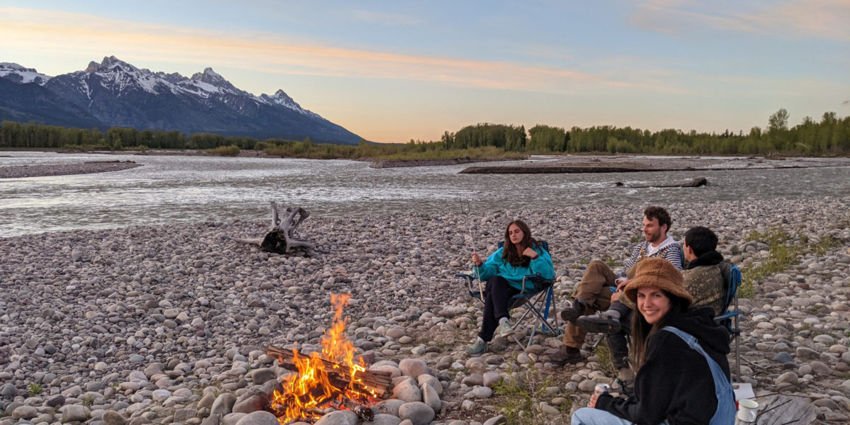 A group of friends sit around a sunset bonfire next to the river and mountains