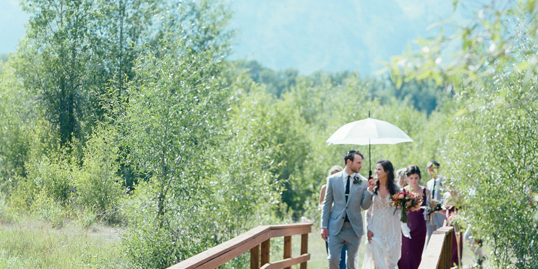 A bride and groom walk across a bridge with an umbrella in front of their guests