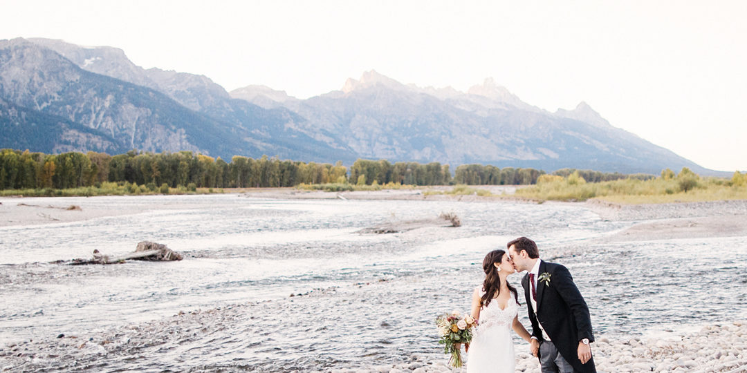 A bride and groom kissing next to a river in front of mountains during the fall