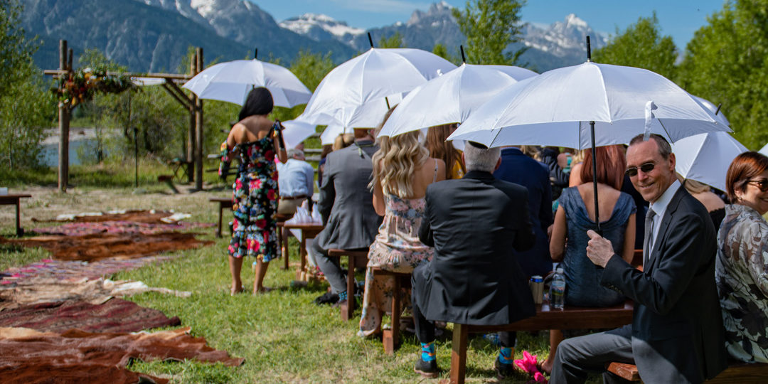 Wedding ceremony guests hold umbrellas on a sunny day in front of mountains